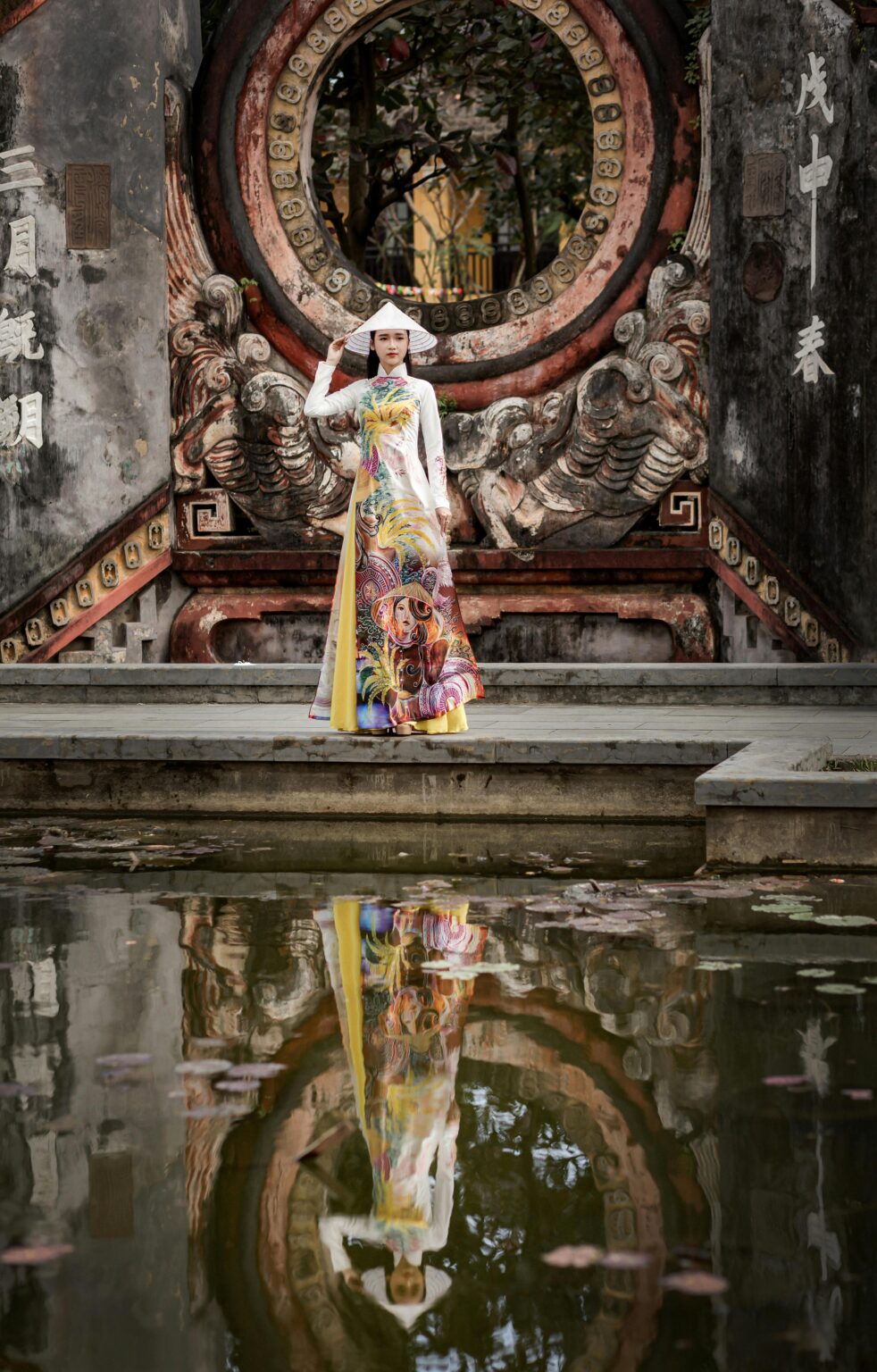 Elegant Vietnamese woman in ao dai stands by a temple with a serene water reflection.