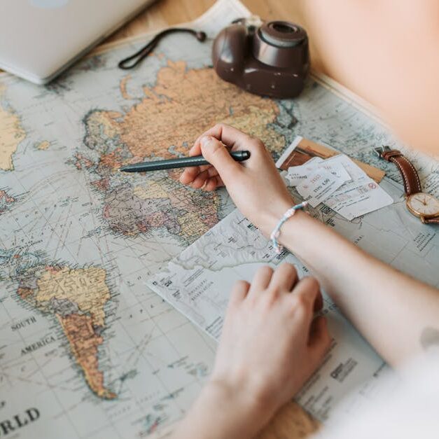 Hands marking destinations on a world map surrounded by travel essentials like passport and camera.